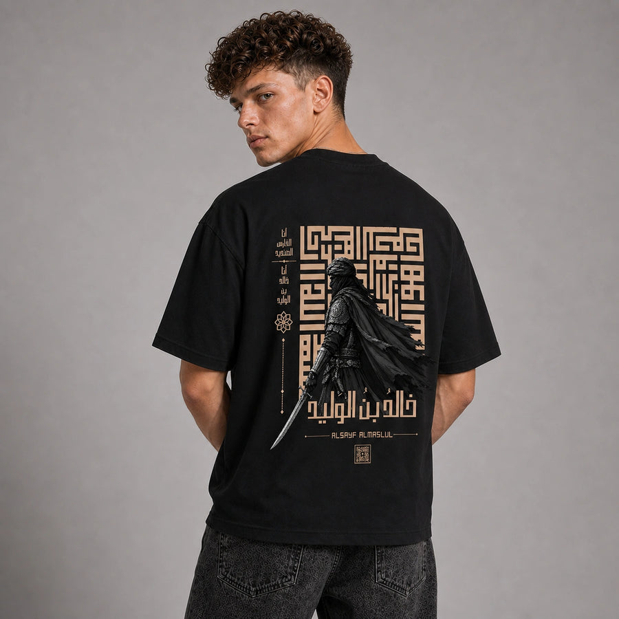 A man wearing a black t-shirt with a large graphic print on the back, featuring a dark knight in a flowing cape and Arabic calligraphy, photographed against a neutral gray studio background.