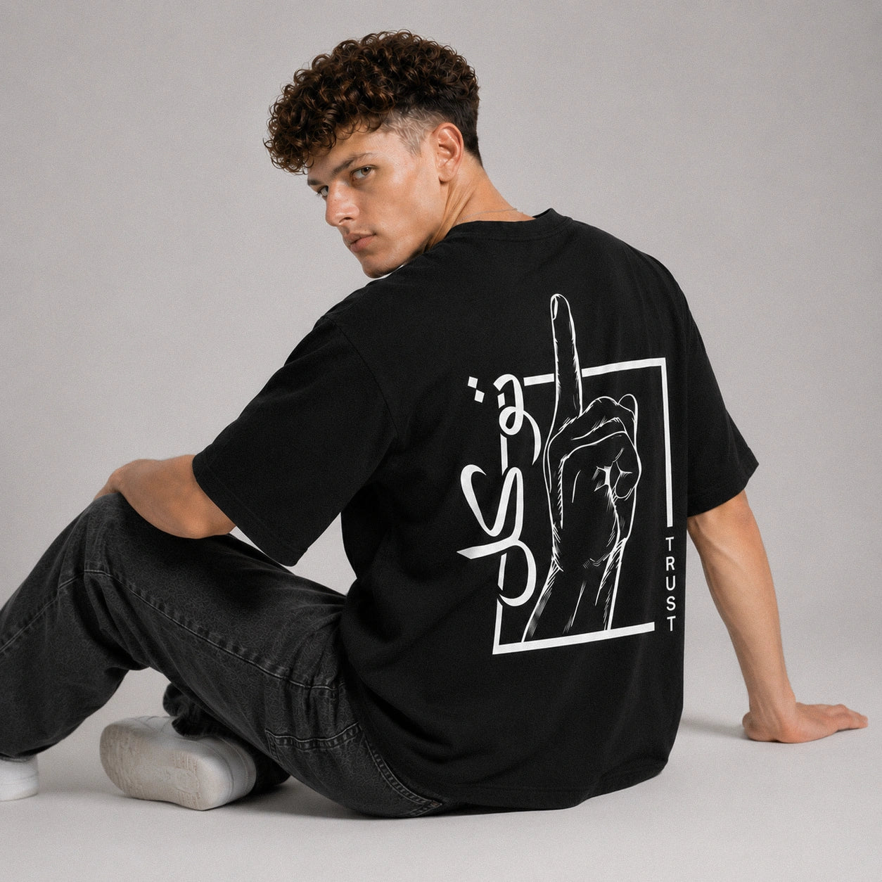 Person wearing a black t-shirt with a graphic design on the back, sitting against a gray background.