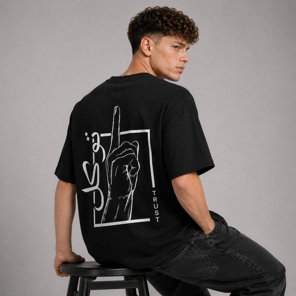 Person wearing a black t-shirt with a graphic design on the back, sitting on a stool against a gray background.