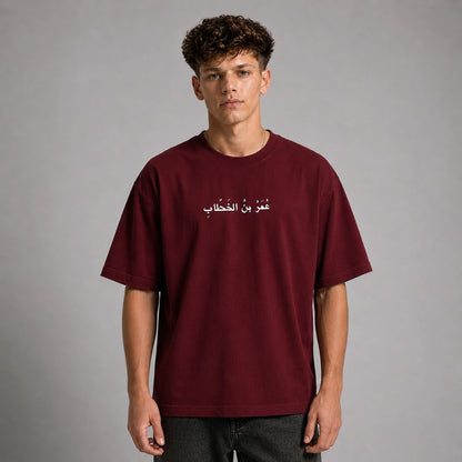 A man wearing a maroon short-sleeve t-shirt with white Arabic text printed on the chest, photographed against a neutral gray studio background.