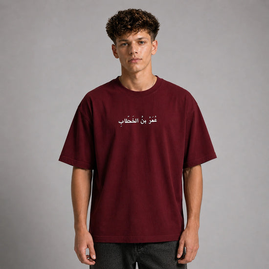 A man wearing a maroon short-sleeve t-shirt with white Arabic text printed on the chest, photographed against a neutral gray studio background.