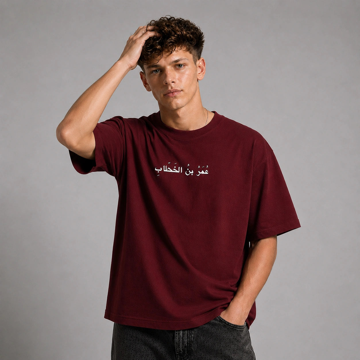 A young man wearing an oversized maroon t-shirt with white Arabic text on the chest, posed against a neutral gray studio background.