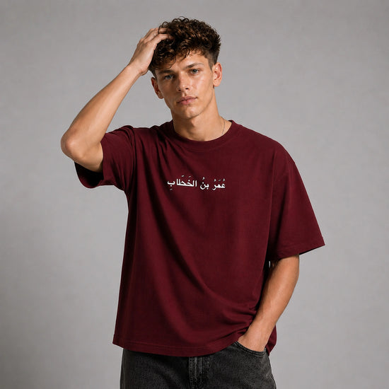 A young man wearing an oversized maroon t-shirt with white Arabic text on the chest, posed against a neutral gray studio background.