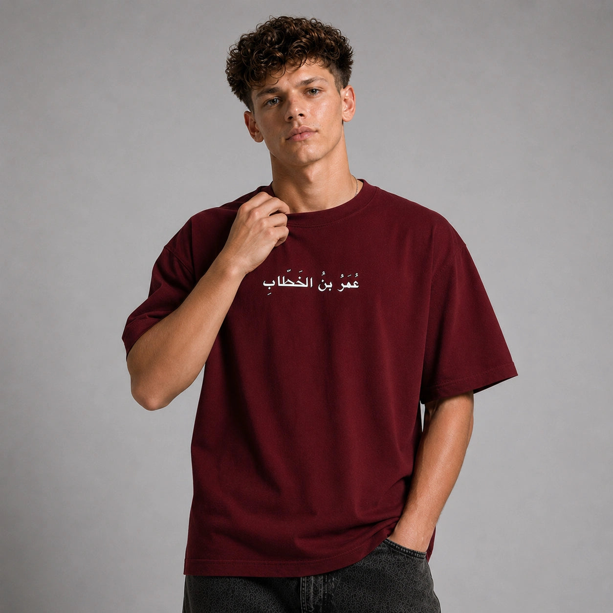 A young man with curly brown hair wearing an oversized maroon t-shirt with white Arabic text across the chest, paired with dark gray jeans, set against a solid gray studio background.