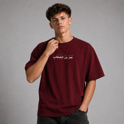 A young man with curly brown hair wearing an oversized maroon t-shirt with white Arabic text across the chest, paired with dark gray jeans, set against a solid gray studio background.