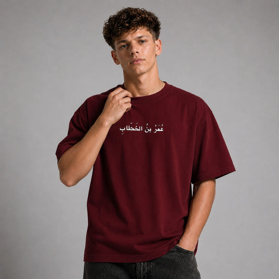 A young man with curly brown hair wearing an oversized maroon t-shirt with white Arabic text across the chest, paired with dark gray jeans, set against a solid gray studio background.