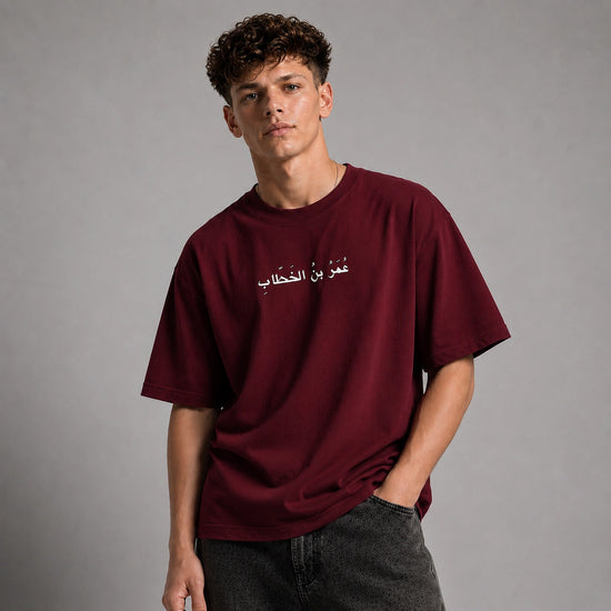 A young man wearing a maroon crewneck t-shirt with white Arabic text across the chest, paired with dark gray jeans, standing against a neutral gray studio background.