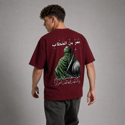 A man wearing a maroon t-shirt from the back, featuring a graphic print of a green-robed warrior with a sword and shield and Arabic text. He is standing against a neutral gray studio background.