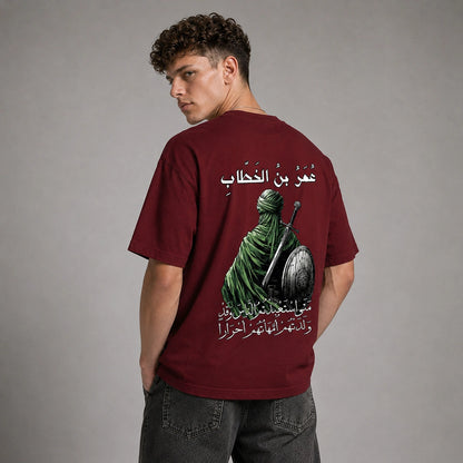 A man wearing a maroon short-sleeve t-shirt with a large graphic print on the back, featuring a green-robed figure and Arabic text, photographed against a neutral gray studio background.