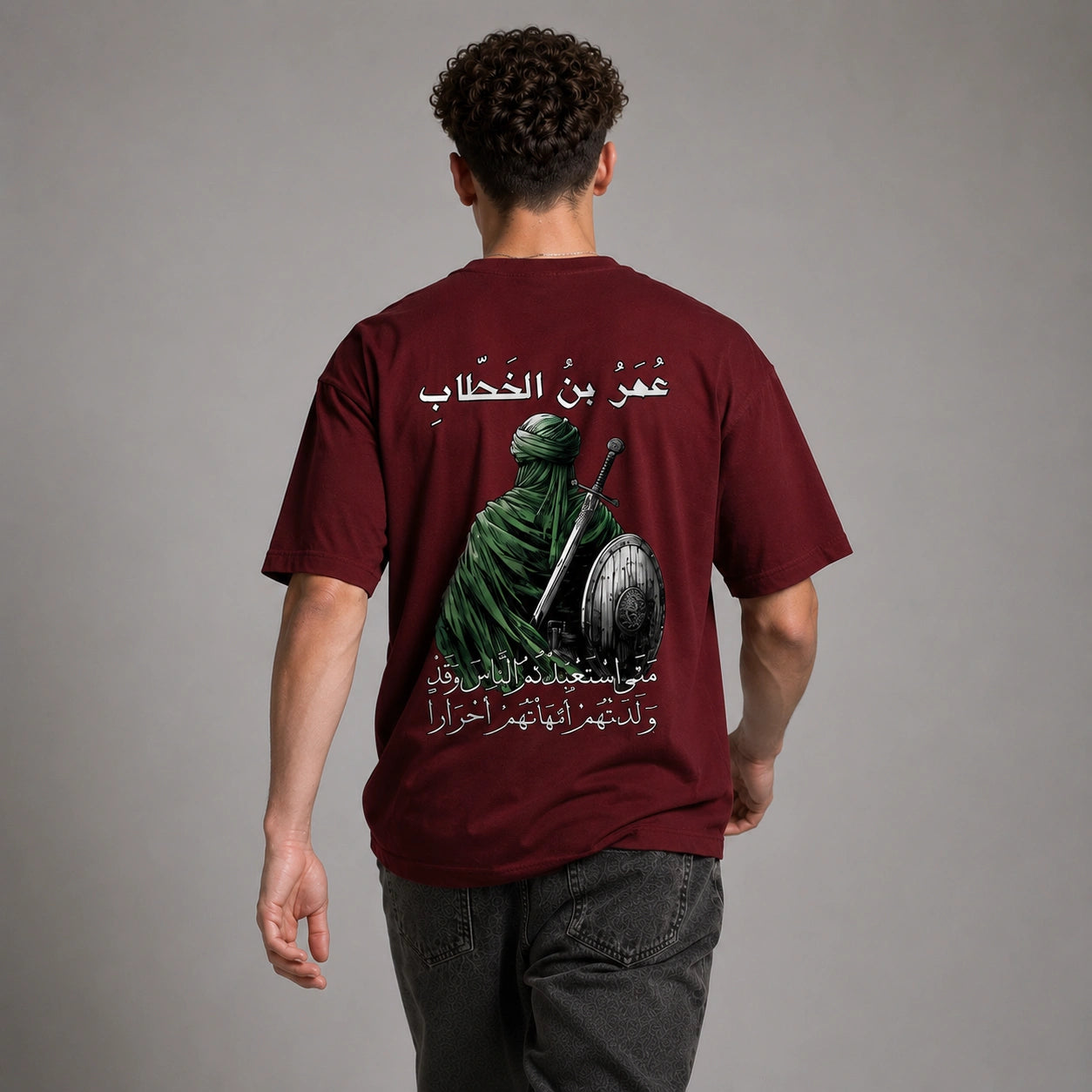 A man viewed from behind wearing a maroon short-sleeve t-shirt with a large graphic print on the back. The print features a green-robed figure holding a sword and shield, with white Arabic text above and below.