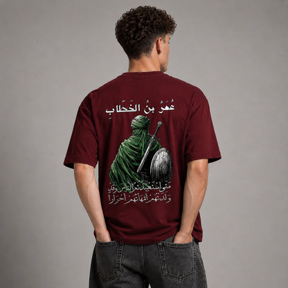 A man viewed from behind wearing a maroon short-sleeve t-shirt with a large graphic print on the back. The print features a green-robed figure holding a sword and shield, with white Arabic text above and below.