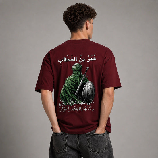 A man viewed from behind wearing a maroon short-sleeve t-shirt with a large graphic print on the back. The print features a green-robed figure holding a sword and shield, with white Arabic text above and below.