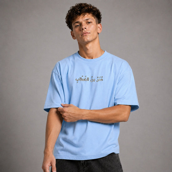 A young man with curly brown hair wearing a light blue crewneck t-shirt with gold Arabic calligraphy on the chest, posed against a neutral gray studio background.