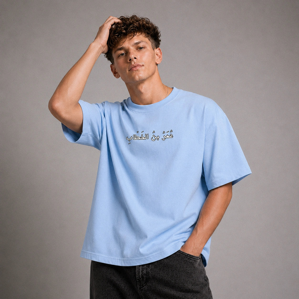 A young man with curly brown hair wearing a light blue crewneck t-shirt with Arabic calligraphy printed across the chest, paired with dark gray jeans, posing against a neutral gray studio background.