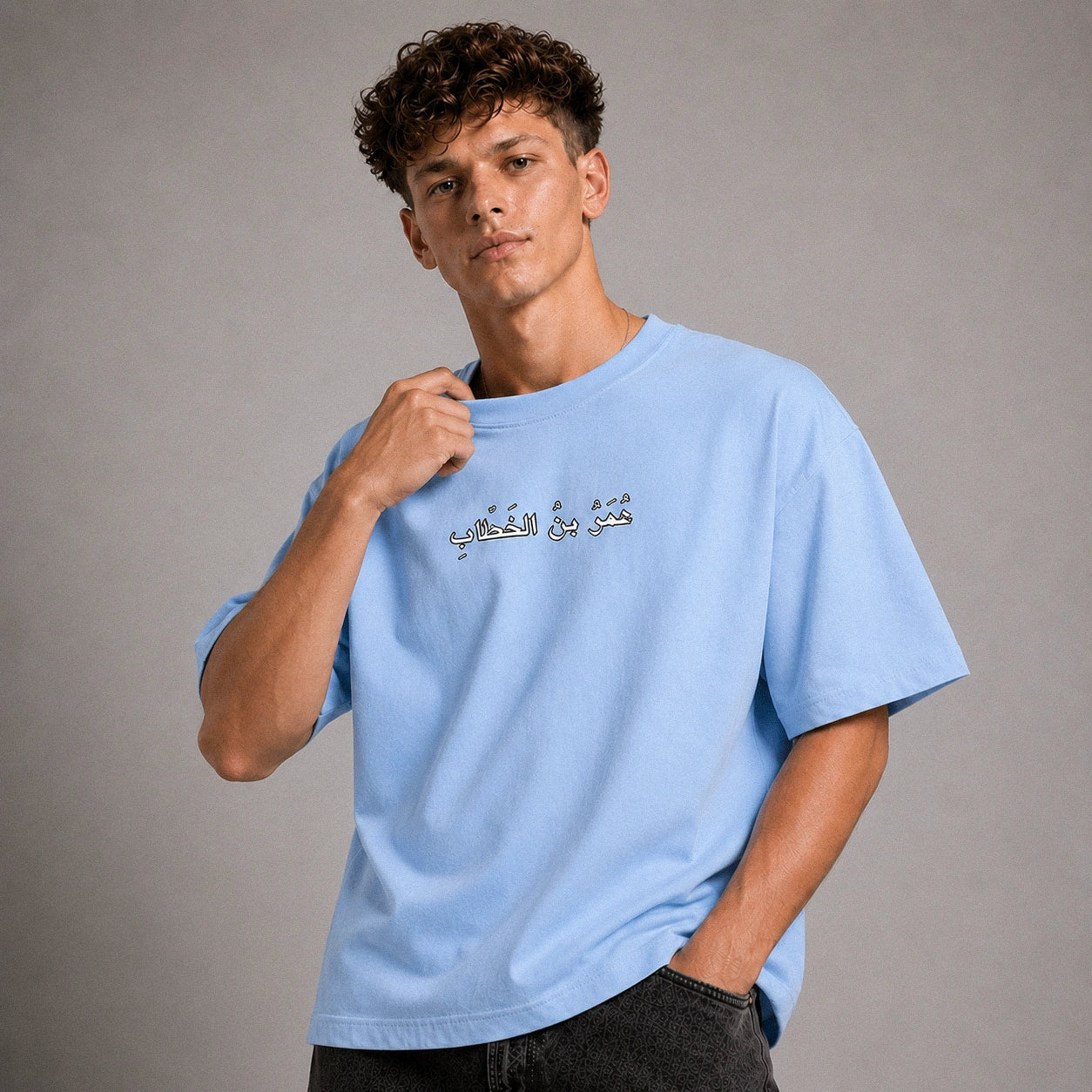 A young man with curly brown hair wearing a light blue crewneck t-shirt with Arabic text printed on the chest, posed against a neutral gray studio background.