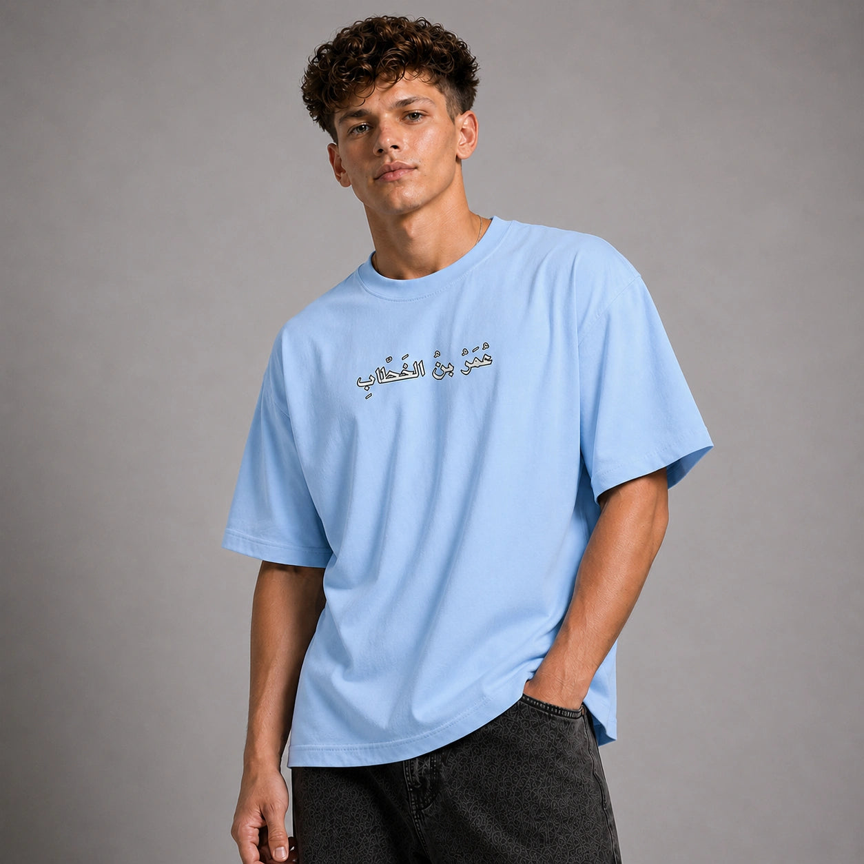 A young man wearing a light blue crewneck t-shirt with Arabic calligraphy printed across the chest, paired with dark gray jeans, photographed against a neutral gray studio background.