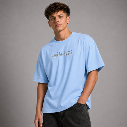 A young man wearing a light blue crewneck t-shirt with Arabic calligraphy printed across the chest, paired with dark gray jeans, photographed against a neutral gray studio background.