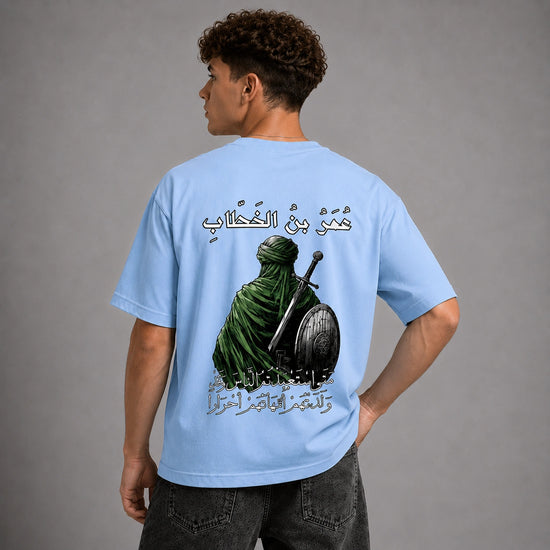 A man shown from the back wearing a light blue t-shirt with a large graphic print of a green-robed warrior holding a sword and shield, accompanied by Arabic text.