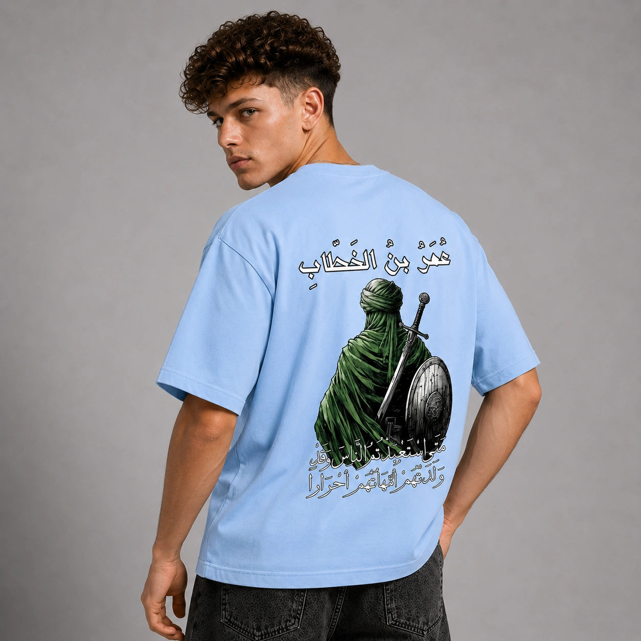 A man wearing a light blue t-shirt with a large graphic print on the back, featuring Arabic calligraphy and an illustration of a robed figure holding a sword and shield. The model is shown from the back against a solid gray studio background.