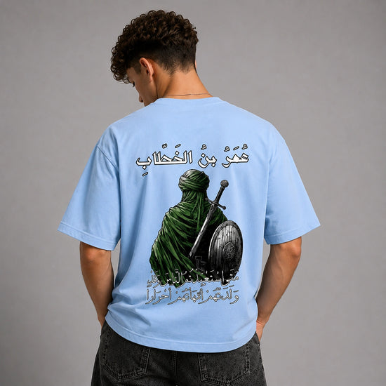 A man viewed from behind wearing a light blue t-shirt with a large graphic print on the back. The graphic features a green-robed figure holding a sword and shield, with Arabic calligraphy above and below.