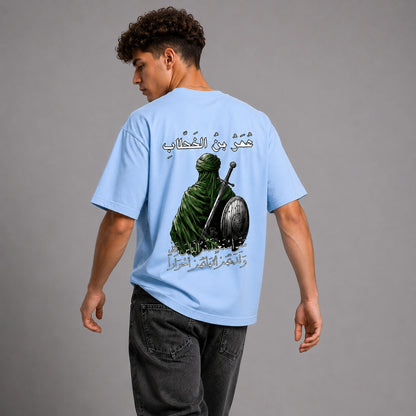 A man wearing a light blue graphic t-shirt, viewed from behind. The shirt features a large graphic print of a green-robed figure with a sword and shield, flanked by Arabic calligraphy.