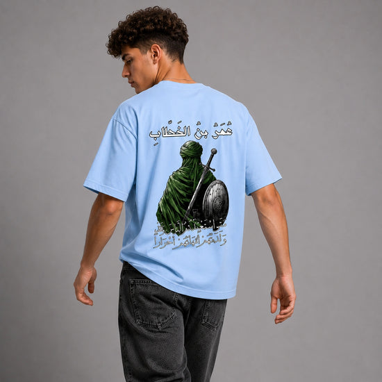 A man wearing a light blue graphic t-shirt, viewed from behind. The shirt features a large graphic print of a green-robed figure with a sword and shield, flanked by Arabic calligraphy.