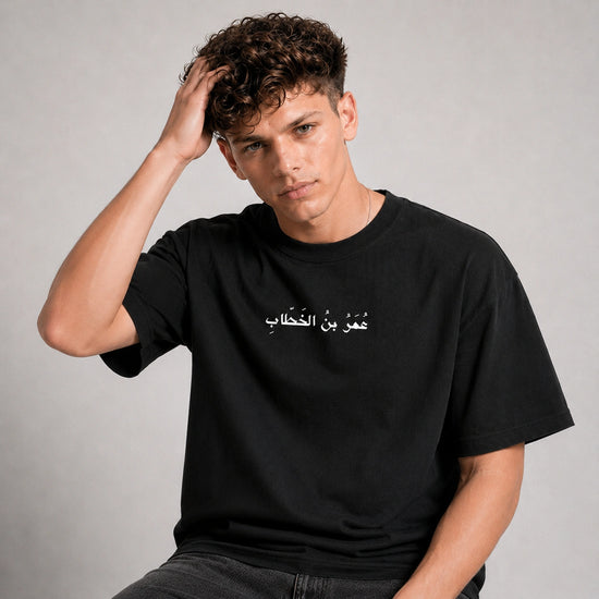 A young man with curly brown hair wearing a black t-shirt with white Arabic text across the chest, posing against a neutral gray studio background.