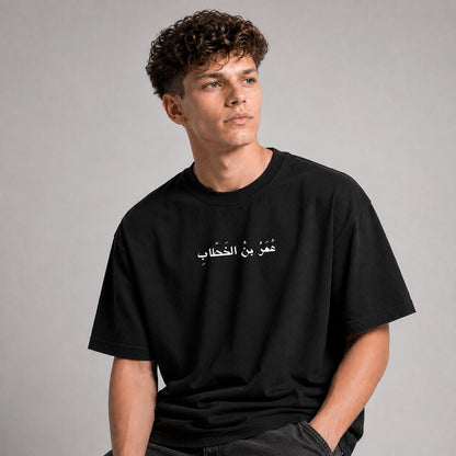 A man wearing a black crew-neck t-shirt with white Arabic text printed across the chest, posed against a neutral gray studio background.