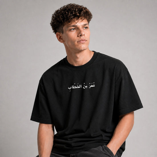 A man wearing a black crew-neck t-shirt with white Arabic text printed across the chest, posed against a neutral gray studio background.