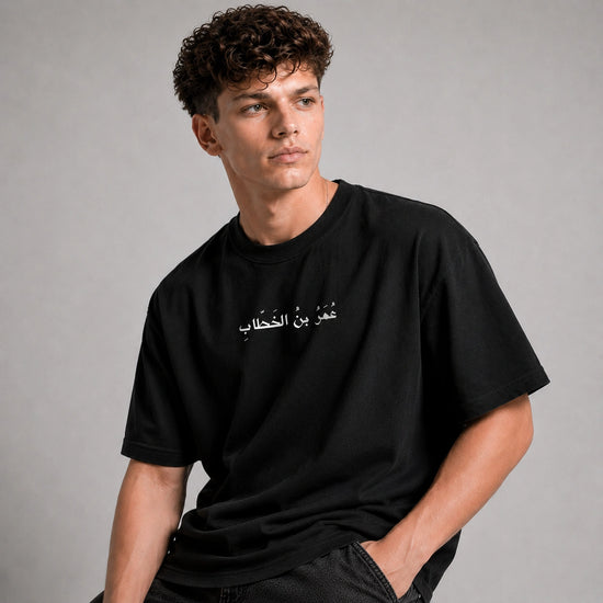 A man wearing a black short-sleeve t-shirt with white Arabic text printed on the chest, posed against a neutral gray studio background.