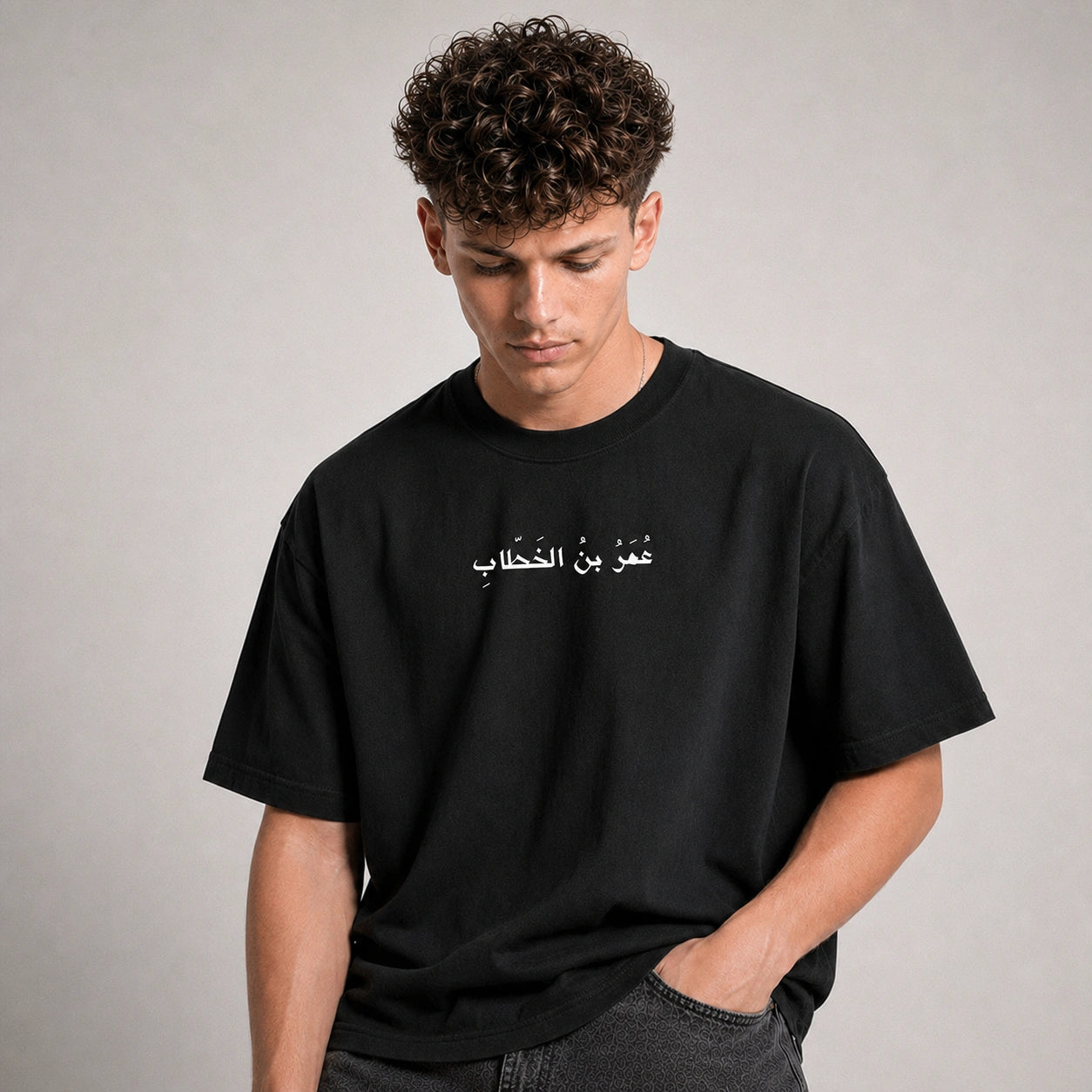 A man wearing a black crewneck t-shirt with white Arabic text printed across the chest, posed against a neutral gray background.