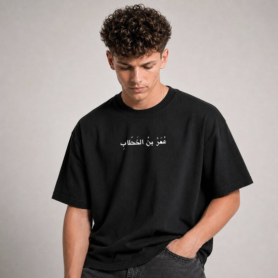 A man wearing a black crewneck t-shirt with white Arabic text printed across the chest, posed against a neutral gray background.