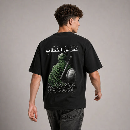 A man viewed from the back wearing a black graphic t-shirt with a large print of a green-robed warrior and Arabic text, paired with dark gray jeans, against a neutral gray studio background.