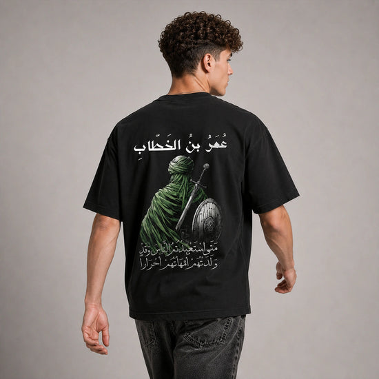 A man viewed from the back wearing a black graphic t-shirt with a large print of a green-robed warrior and Arabic text, paired with dark gray jeans, against a neutral gray studio background.