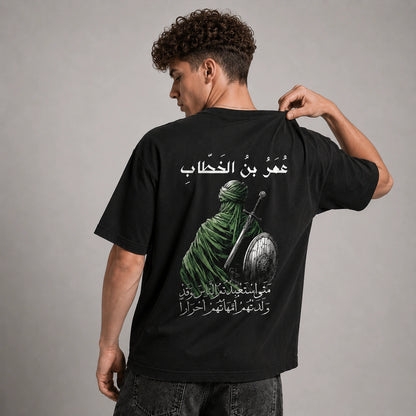 A man wearing a black t-shirt, shown from the back, featuring a graphic print of a green-robed warrior with a sword and shield and white Arabic text.