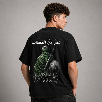 A man shown from the back wearing a black t-shirt with a large graphic print of a green-clad figure holding a sword and shield, accompanied by white Arabic text, against a neutral gray studio background.