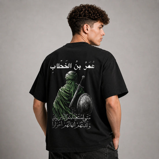 A man shown from the back wearing a black t-shirt with a large graphic print of a green-clad figure holding a sword and shield, accompanied by white Arabic text, against a neutral gray studio background.