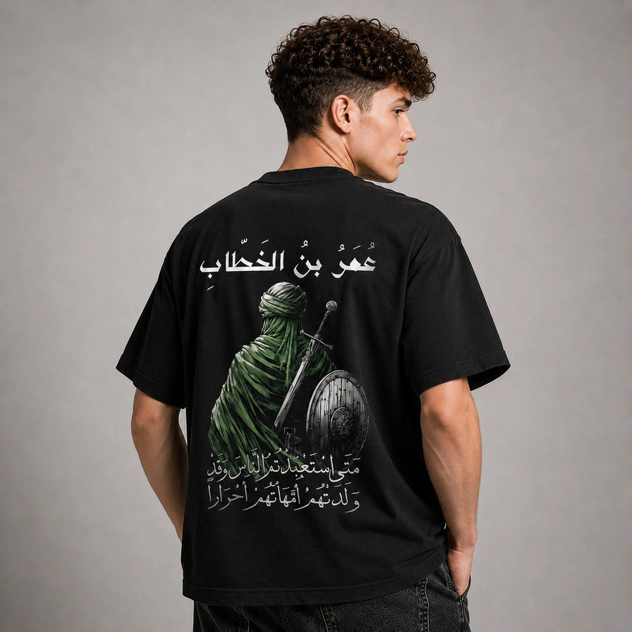 A man shown from the back wearing a black t-shirt with a large graphic print of a green-clad figure holding a sword and shield, accompanied by white Arabic text, against a neutral gray studio background.