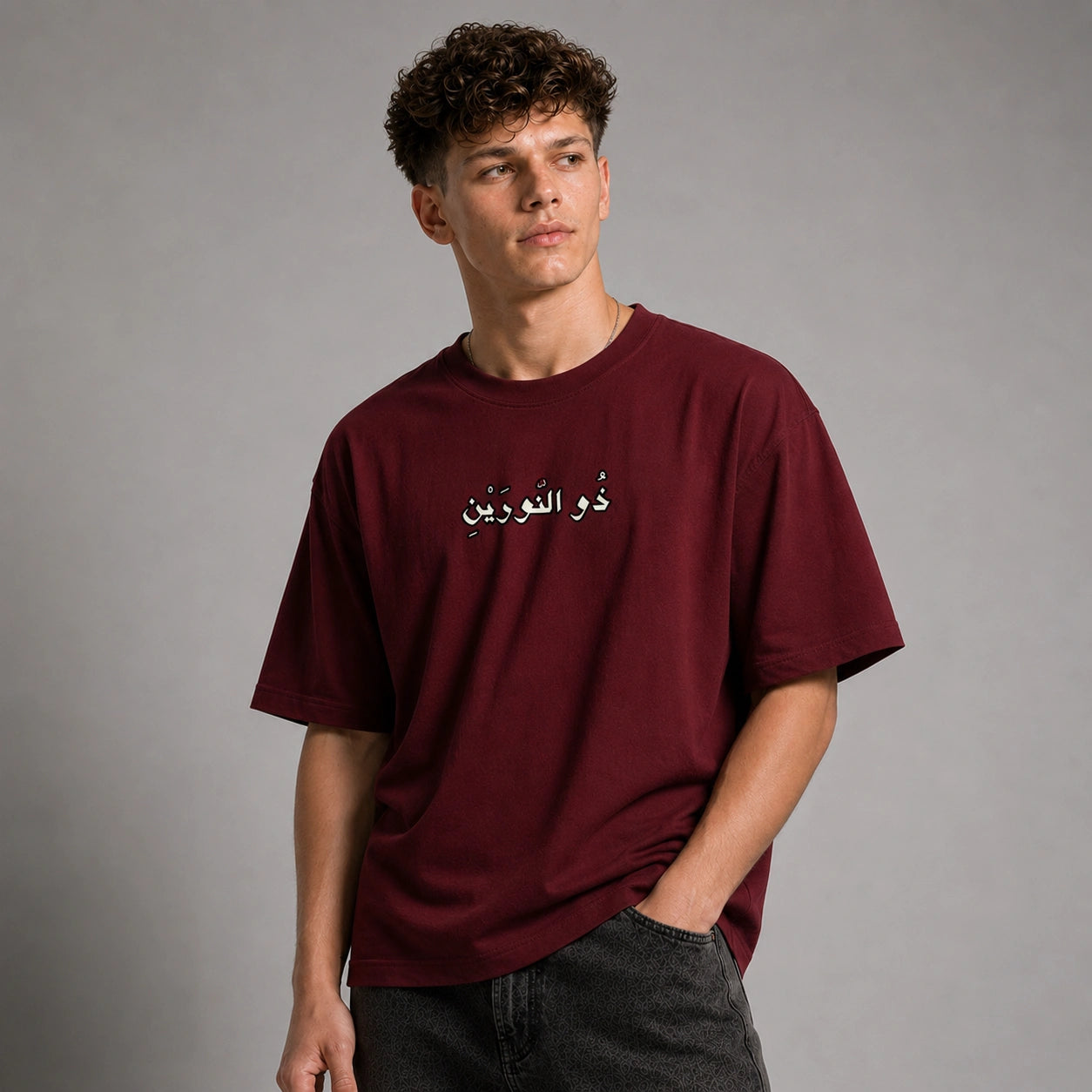 A young man with curly brown hair wearing an oversized maroon t-shirt with white Arabic script on the chest, paired with dark gray jeans, standing against a neutral gray studio background.