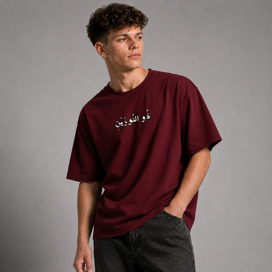 A young man with curly brown hair wearing a maroon short-sleeve t-shirt with white Arabic text on the chest, paired with dark gray jeans, standing against a solid gray studio background.