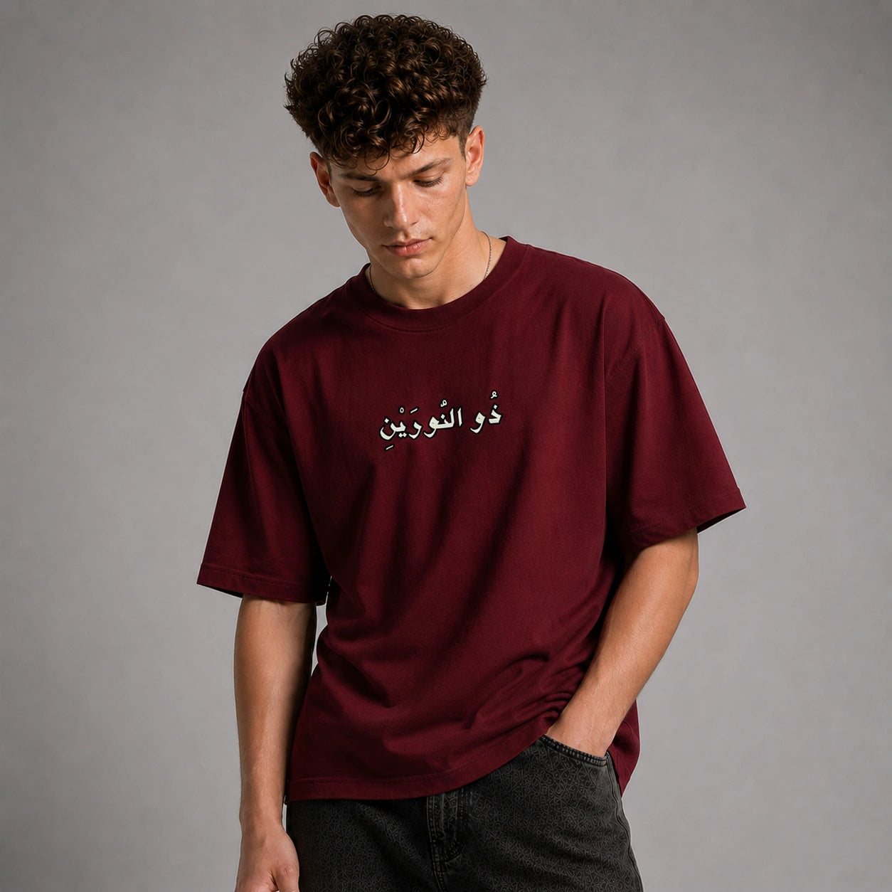 A young man with curly brown hair wearing an oversized maroon t-shirt with white Arabic text on the chest, paired with dark gray jeans, standing against a solid gray studio background.