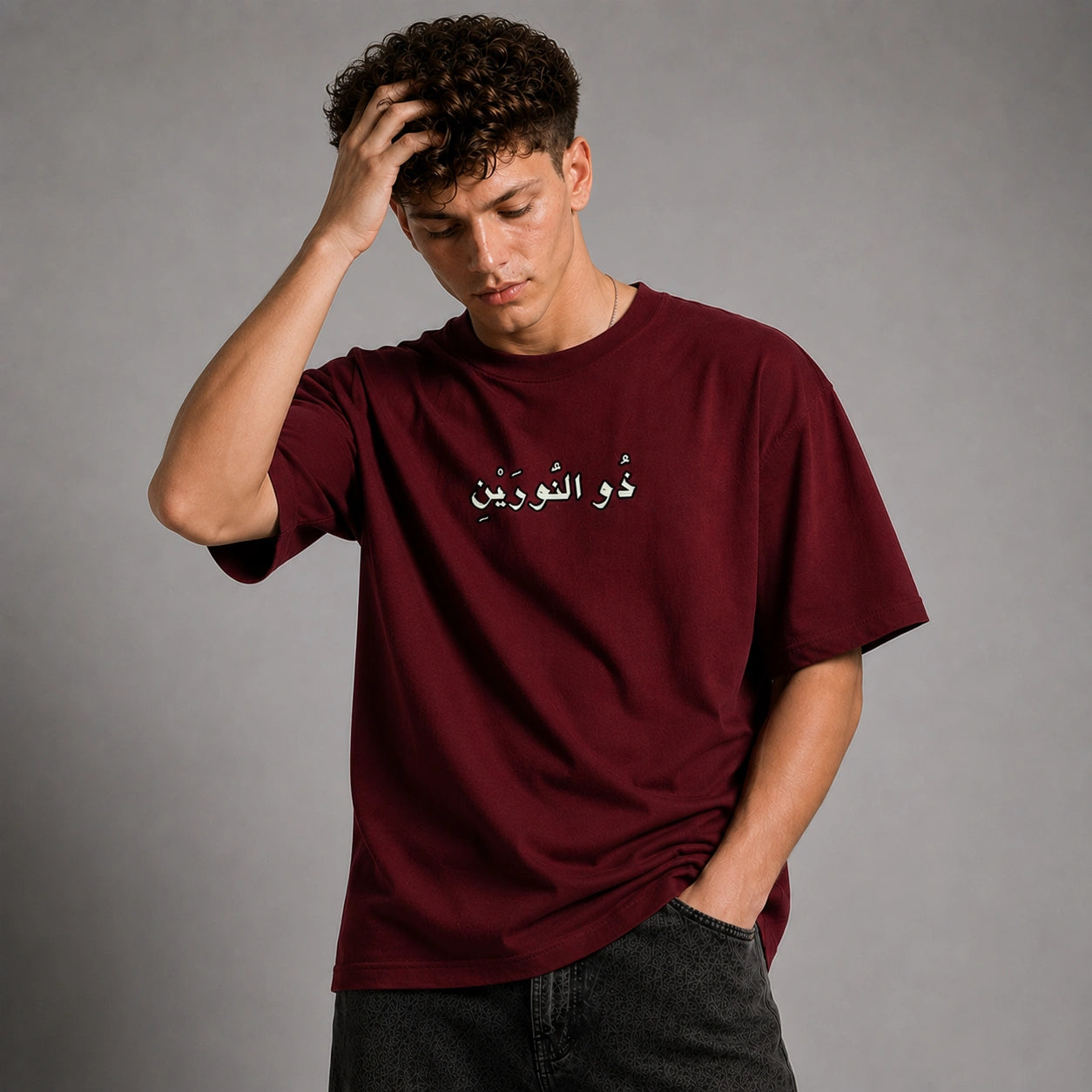 A man wearing a maroon crewneck t-shirt with white Arabic text on the chest, paired with dark gray jeans, posing against a neutral gray background.