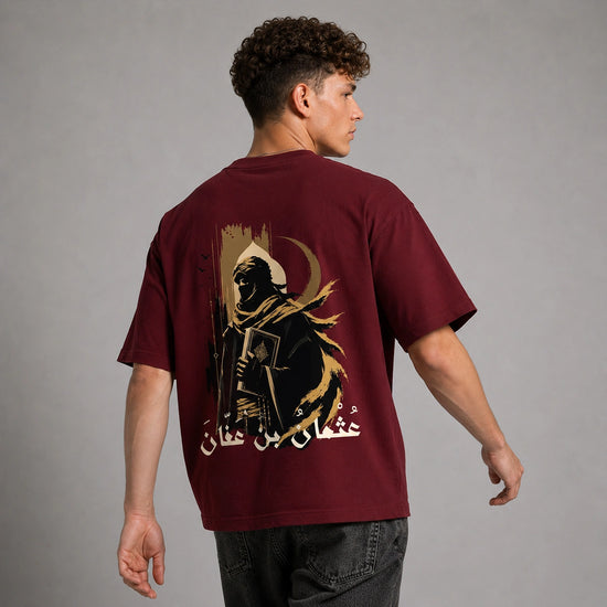 A man wearing an oversized maroon t-shirt, viewed from the back. The shirt features a large graphic print of a hooded figure holding a book, a crescent moon, and Arabic calligraphy in black and gold.