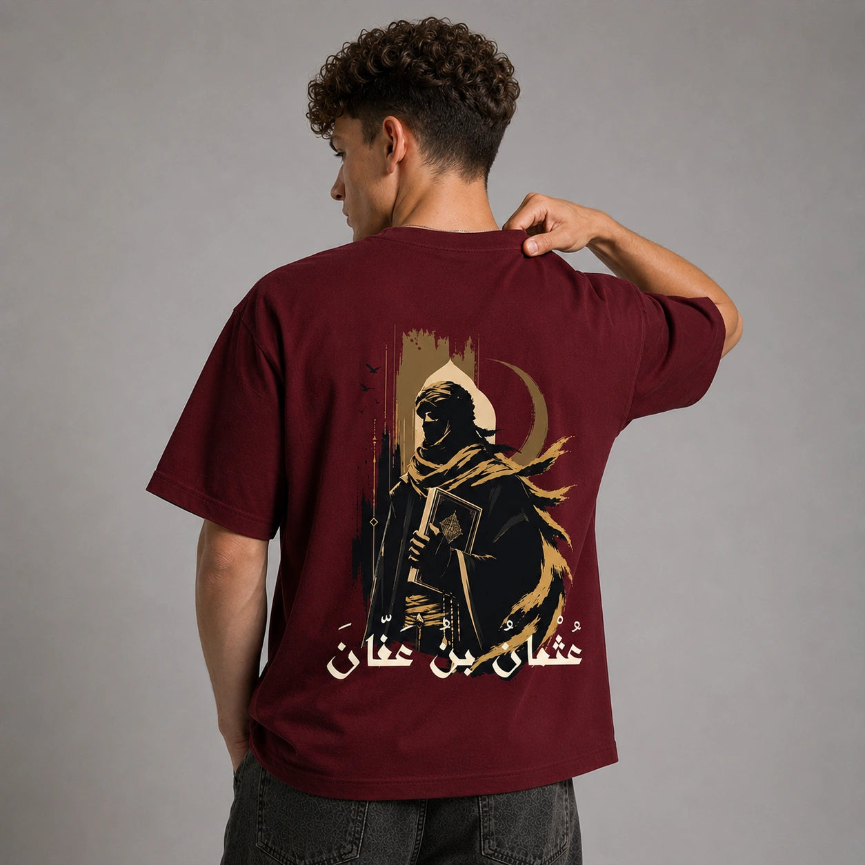 A man shown from behind wearing a maroon short-sleeve t-shirt with a large graphic print on the back. The graphic features a hooded figure in black and gold, a crescent moon, and Arabic calligraphy.