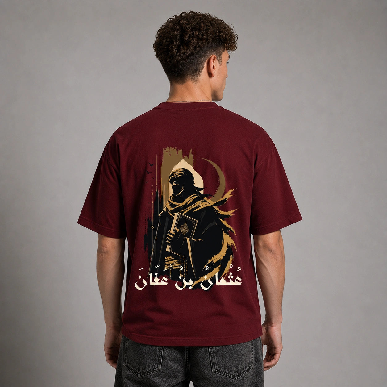 A man viewed from the back wearing a maroon short-sleeve t-shirt with a large graphic print of a hooded figure and Arabic text on the back, paired with dark gray jeans against a neutral gray studio background.