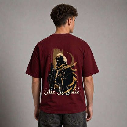 A man viewed from the back wearing a maroon short-sleeve t-shirt with a large graphic print of a hooded figure and Arabic text on the back, paired with dark gray jeans against a neutral gray studio background.