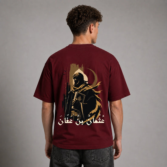 A man viewed from the back wearing a maroon short-sleeve t-shirt with a large graphic print of a hooded figure and Arabic text on the back, paired with dark gray jeans against a neutral gray studio background.