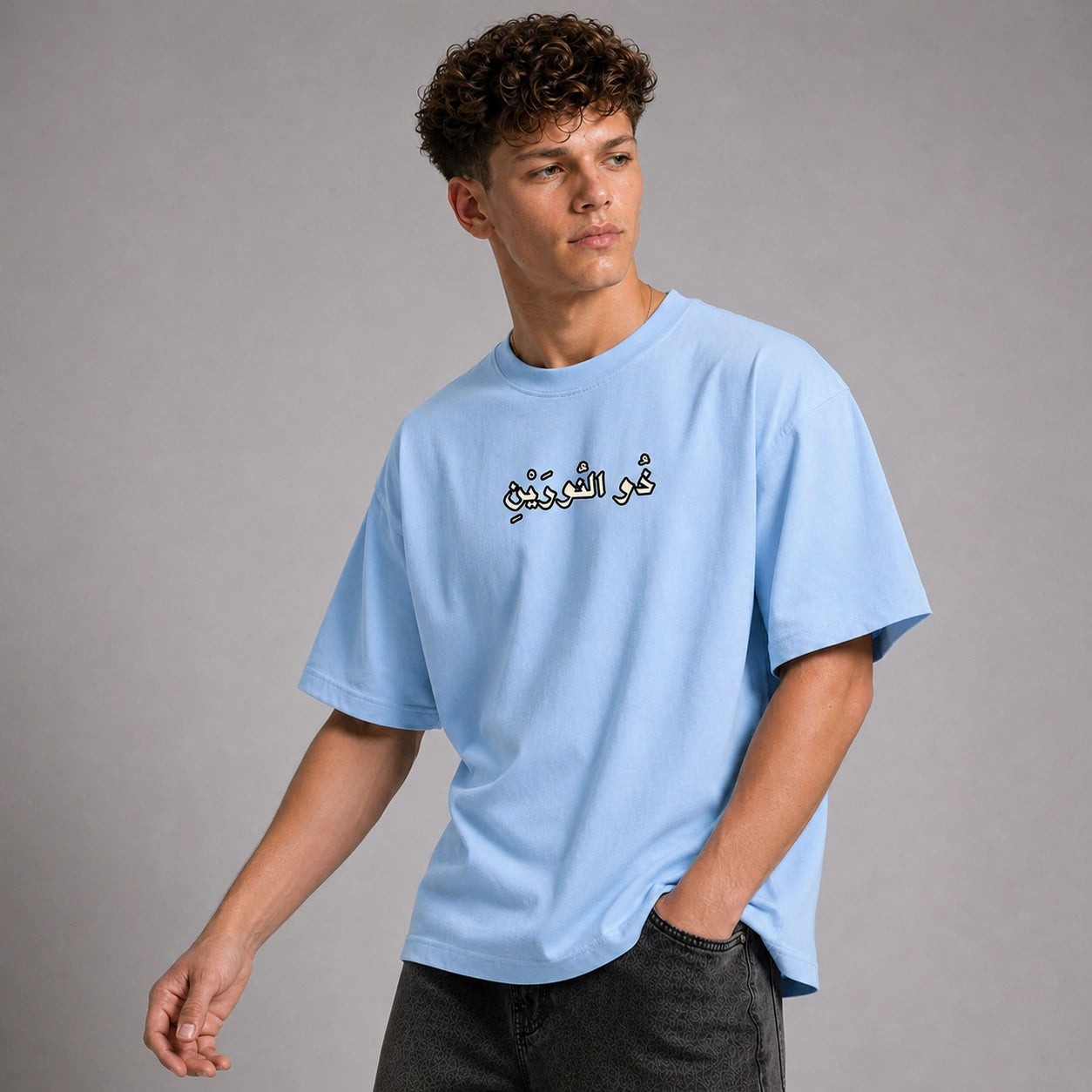 A young man wearing a light blue crewneck t-shirt with black Arabic calligraphy on the chest, paired with dark gray jeans, photographed against a neutral gray studio background.