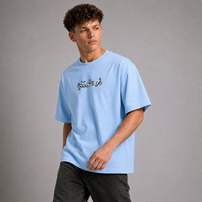 A young man wearing a light blue t-shirt with black Arabic calligraphy on the chest, paired with dark gray jeans, standing against a neutral gray studio background.