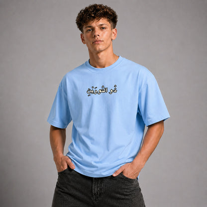 A young man wearing a light blue crewneck t-shirt with black Arabic calligraphy on the chest, paired with dark gray jeans, standing against a solid gray studio background.